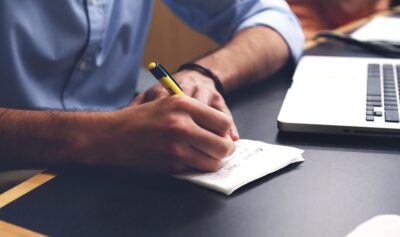 Personne écrivant à la main sur un bloc-notes à côté d'un ordinateur portable sur un bureau
