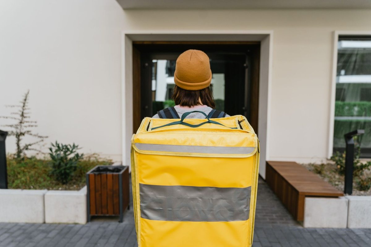 Individu avec sac jaune devant une porte d'immeuble, végétation et banc à proximité