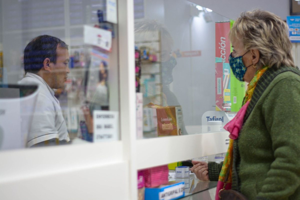 Femme en masque vert et homme en blouse dans une pharmacie, échangeant derrière une vitre