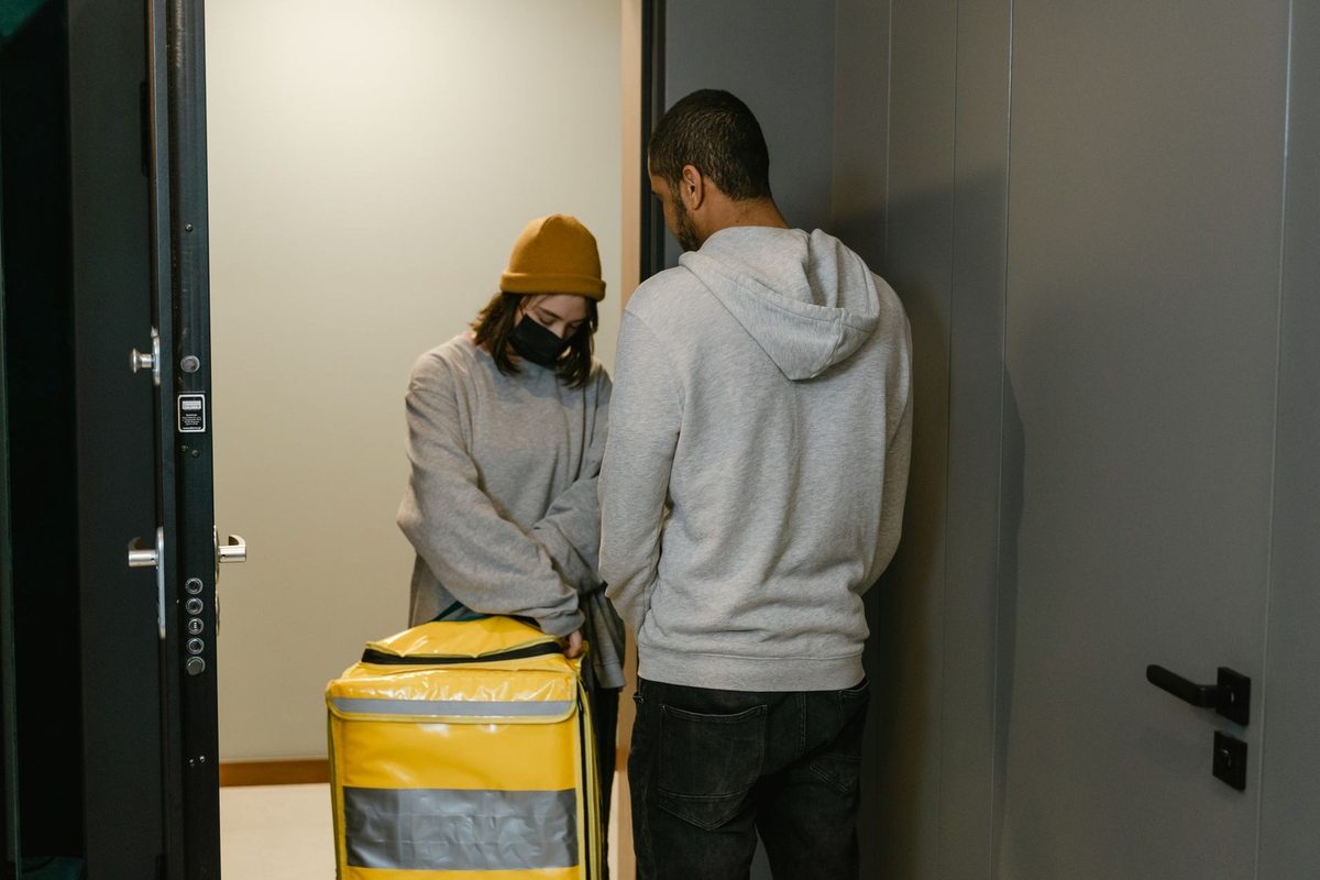 Deux individus devant une porte, l'un portant un sac jaune avec une étiquette, l'autre observant